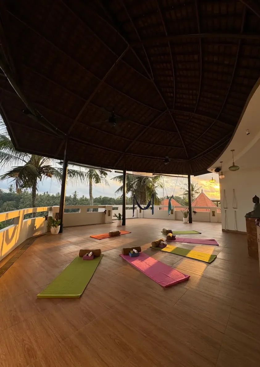 The open-air rooftop yoga pavilion at Bodhi Leaf Wellness Resort at sunset: a thatched roof over a wooden floor with five coloured mats and bolsters laid out, palms and a Balinese temple roof beyond the balcony, and a small Buddha statue by the entrance.