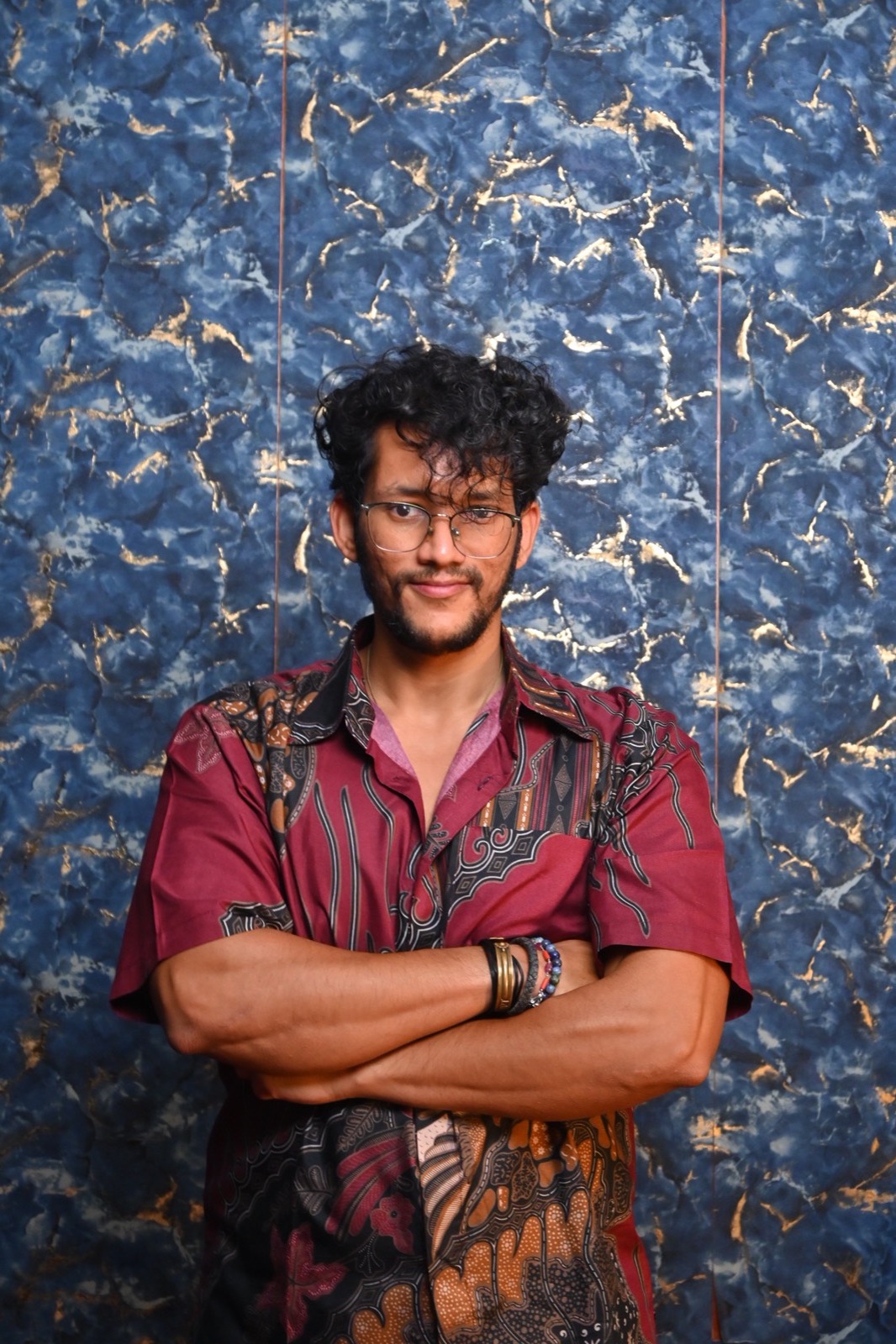 Dushyant Dharma standing with arms crossed in a maroon batik shirt, against a blue and gold marbled wall.
